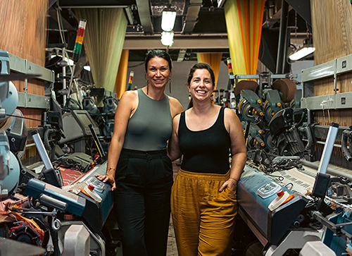 Mélanie Bernard et Trisha Pitura posent au milieu de l’atelier de production de MINI TIPI, avec des métiers à tisser industriels de part et d’autres de la photo.